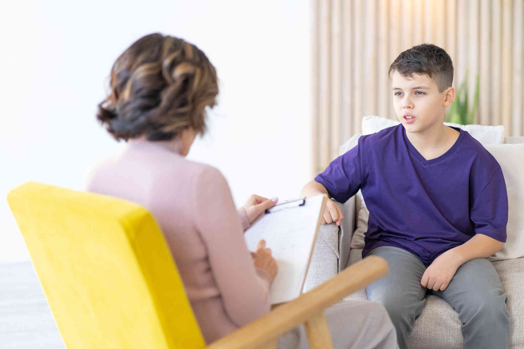 A boy in a purple shirt sits on a couch while talking to the female therapist sitting across from him.